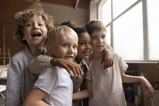 Four multiethnic children laughing staring at camera, standing, hugging in creative workshop after painting class. Portrait of four carefree and happy boys and African girl embracing enjoy friendship