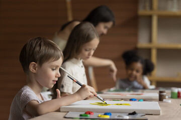 Pretty pupil boy painting during group class, studying in art-school for little children, drawing pictures on canvas sit at table, holding paintbrush, using palette with paints, attend creative course