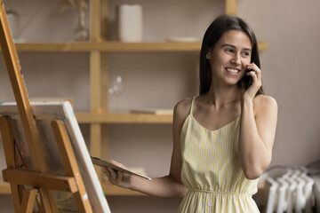 Attractive female painter, art-school student talking on smartphone standing in cozy workshop, lead pleasant personal conversation, distracted from painting on easel, looks satisfied chatting indoors