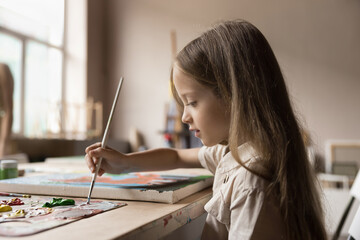 Cute serious little girl painting pictures sit at table in workshop, child holding paintbrush drawing with paints on canvas looking concentrated, enjoy creative hobby in studio. Development, vocation - obrazy, fototapety, plakaty