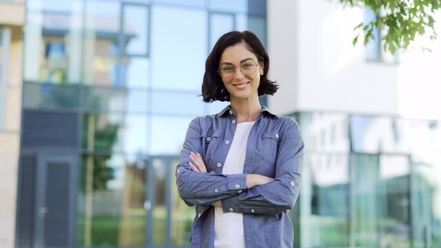 Portrait of smiling female student looking at camera while standing in campus space near university building. Head shot of a positive brunette teacher in glasses and a shirt posing with crossed arms