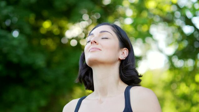Close up. Happy young cute woman relaxing with closed eyes standing in urban city park. A smiling beautiful fit female brunette breathes deeply and enjoys being in nature looking at the camera