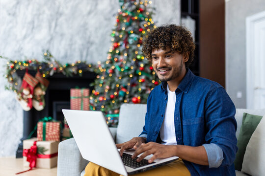 Young Smiling Man Working Remotely From Home For Christmas Sitting On Couch In Living Room Near Decorated Christmas Tree, Hispanic Man Using Laptop, Typing On Keyboard.