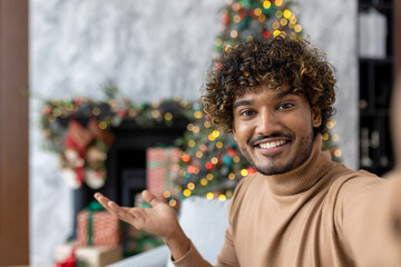 Online video call, remote conversation for Christmas, man uses app on phone to communicate with friends, records greetings and invitations for New Year's celebration, sits on sofa and looks camera.