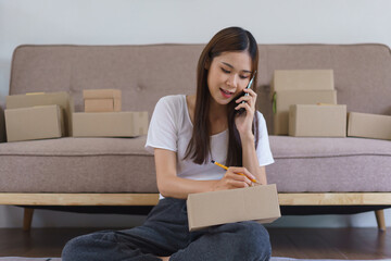 Online business concept, Asian business woman talks on smartphone and writes address on parcel box