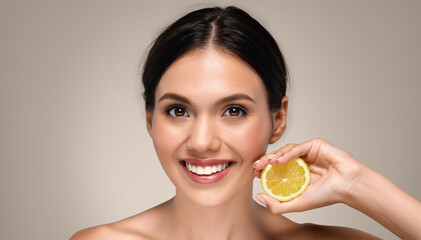 Smiling young european woman with perfect face, show yellow lemon fruit, isolated on gray studio background