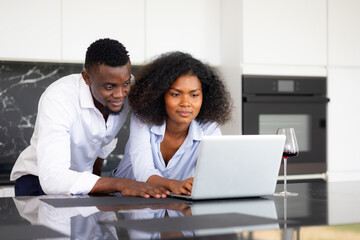 Happy excited successful African American man and woman couple togetherness at home office. Work from home. Couple using laptop computer in kitchen at home