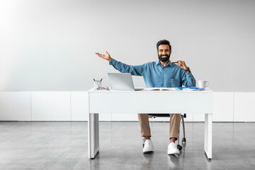 Handsome indian businessman sitting at desk with laptop and pointing aside at free place with hand