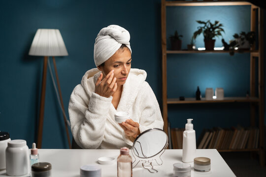 Happy woman with towel on head doing routine skin care at home with beauty products, applying moisturizer on her face near mirror.