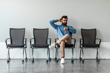 Worried indian job seeker waiting for interview far too long, sitting on chair alone in row,...