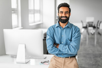 Happy indian businessman standing by his workplace with folded arms, smiling at camera