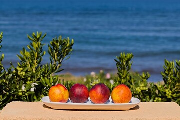 Four ripe peaches align on a white plate by the Sardinian sea. Bathed in southern sun, it's a...