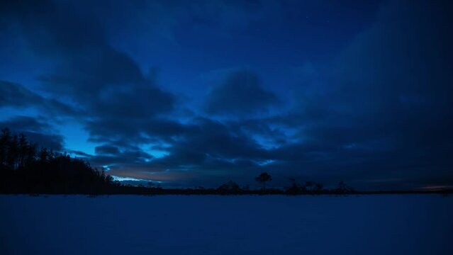 A time lapse of the blue color of evening over snowy marshland with clouds in the starry sky in winter in Finland