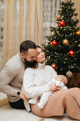 Obraz premium Young parents is holding a sleeping newborn baby in their arms near the Christmas tree at home on Christmas. 
