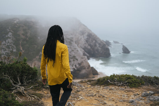 African male wearing long dreadlocks standing on ocean shore, white waves and cliffs in fog on background