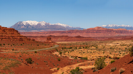 Fototapeta premium Panoramic view of a desert road in southern Utah, USA with snowy mountains in the horizon and clear blue sky.