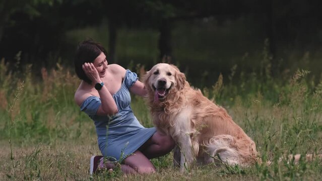 Pretty girl owner hugging golden retriever dog and smiling at nature. Young woman petting purebred pet doggy labrador at summertime