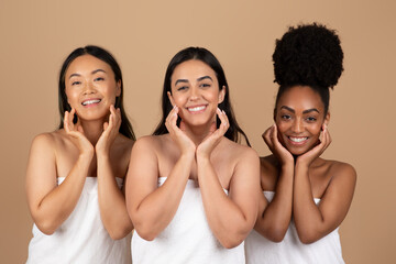 Multiracial women in towels touch cheeks, beige studio background