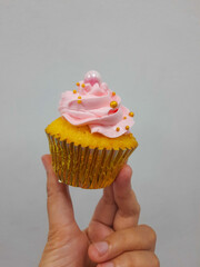 Yellow cupcake with pink cream, edible pearls and gold cupcake liner. Hold by hand, isolated on white background