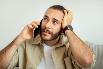 Confused Young Guy Talking On Cellphone And Touching Head