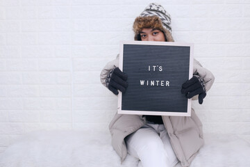 Young Asian woman in winter clothes showing lettering board with text It's Winter 
