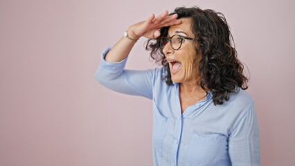 Middle age hispanic woman looking for someone surprised over isolated pink background