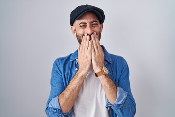 Hispanic man with beard standing over isolated background laughing and embarrassed giggle covering...