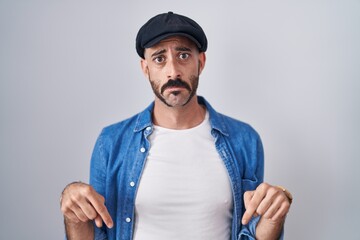 Hispanic man with beard standing over isolated background pointing down looking sad and upset, indicating direction with fingers, unhappy and depressed.