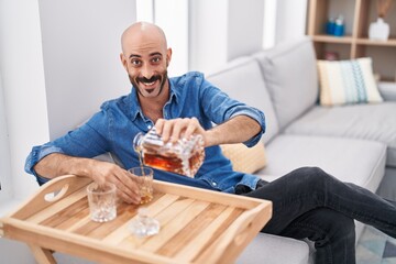 Young hispanic man pouring whisky on glass sitting on sofa at home