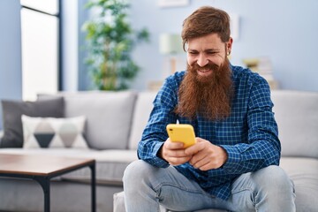 Young redhead man using smartphone sitting on sofa at home