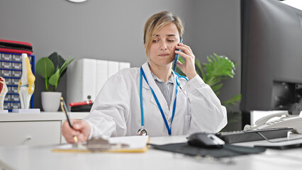 Young blonde woman doctor talking on smartphone writing on document at clinic