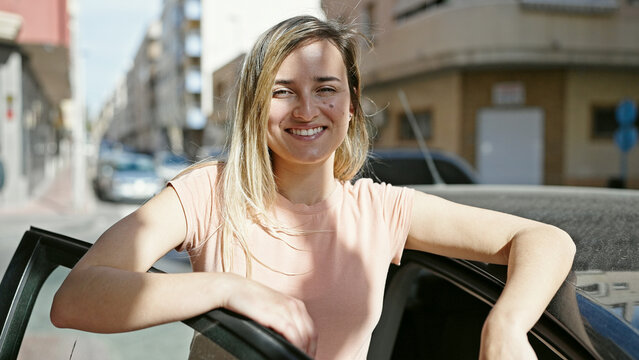 Young blonde woman smiling confident leaning on car door at street