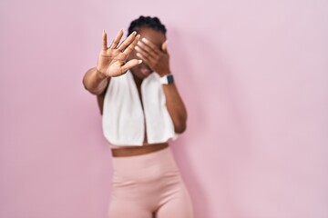 Beautiful black woman wearing sportswear and towel over pink background covering eyes with hands...
