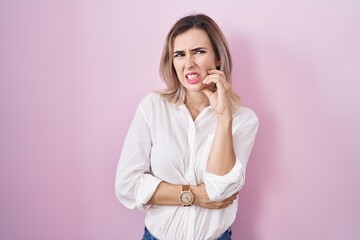 Young beautiful woman standing over pink background looking stressed and nervous with hands on mouth biting nails. anxiety problem.