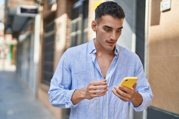 Young hispanic man using smartphone with surprise expression at street