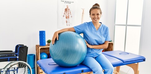 Young beautiful hispanic woman physiotherapist smiling confident holding fit ball at rehab clinic