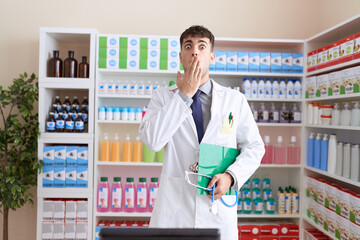 Young hispanic man working at pharmacy drugstore holding stethoscope covering mouth with hand,...