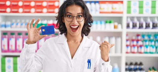 Rolgordijnen Young hispanic woman working at pharmacy drugstore holding credit card pointing thumb up to the side smiling happy with open mouth © Krakenimages.com
