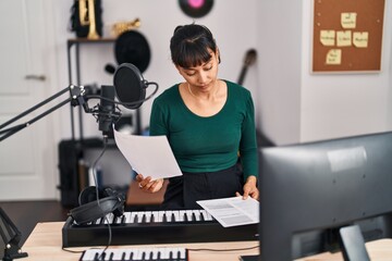 Young beautiful hispanic woman musician reading document sitting on chair at music studio