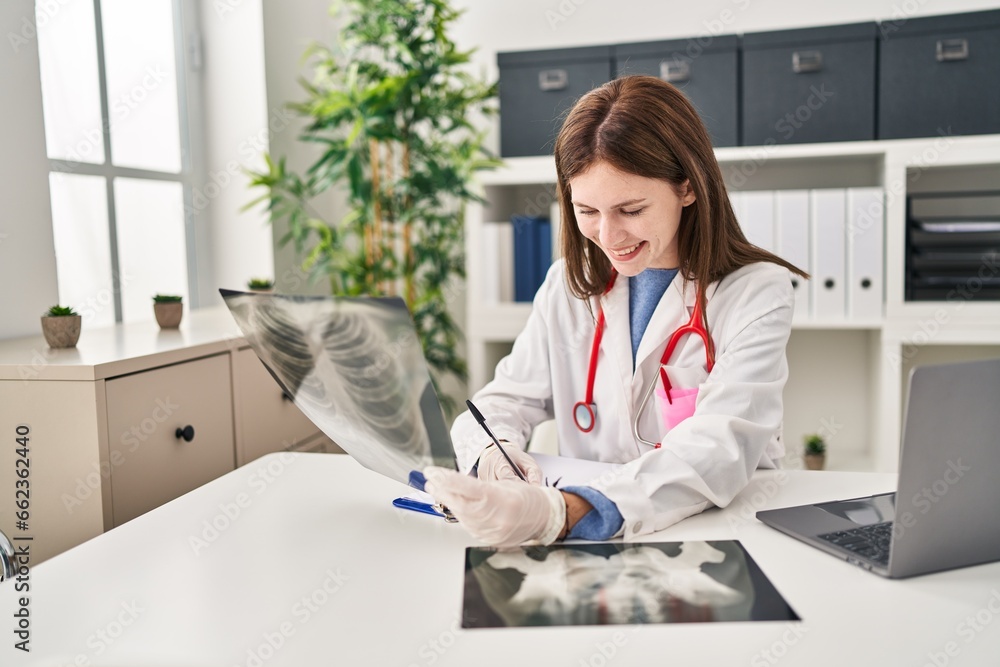 Poster Young blonde woman doctor looking xray writing medical report at clinic