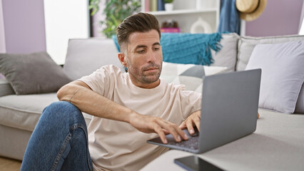 Attractive young hispanic man, concentrated and serious, sitting on the floor in his cozy...