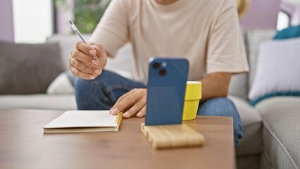 Fototapeta premium Handsome young hispanic man leisurely studies at home, engrossed in writing in his smart notebook, touching smartphone screen, sipping on a cup of coffee whilst sitting on the living room sofa.