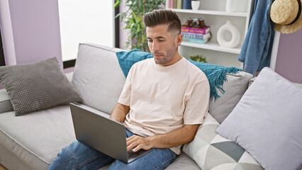 Focused young hispanic man, engrossed in his laptop while working online, sitting, on a cozy sofa...