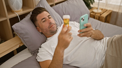 Troubled young hispanic man lying ill in pyjamas on his bed, anxiously looking at his smartphone while clutching a bottle of pills in his bedroom.