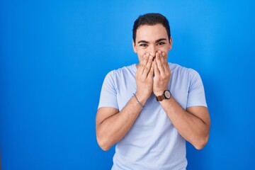 Young hispanic man standing over blue background laughing and embarrassed giggle covering mouth...