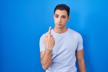 Young hispanic man standing over blue background showing middle finger, impolite and rude fuck off...