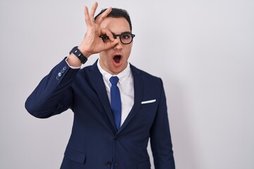 Young hispanic man wearing suit and tie doing ok gesture shocked with surprised face, eye looking through fingers. unbelieving expression.