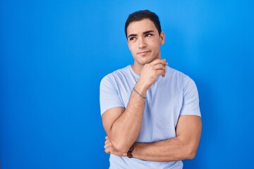 Young hispanic man standing over blue background with hand on chin thinking about question, pensive expression. smiling with thoughtful face. doubt concept.