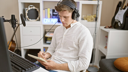 Fototapeta na wymiar Focused young caucasian man, a handsome musician, engrossed in composing an acoustic song at a music studio, listening to melodies, notebook in hand, indoor ambiance