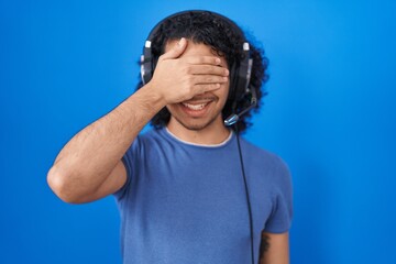 Hispanic man with curly hair listening to music using headphones smiling and laughing with hand on face covering eyes for surprise. blind concept.